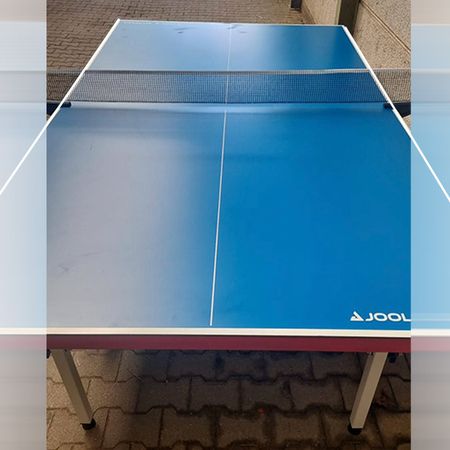 Blaue Tischtennisplatte der Marke Joola mit weißem Rand und Netz in der Mitte, aufgestellt im Freien auf einem gepflasterten Boden zwischen zwei Gebäudewänden.