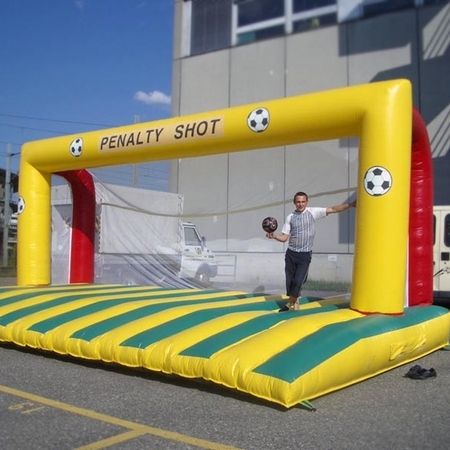 Aufblasbares Fußball-Tor mit gelbem Rahmen und der Aufschrift „Penalty Shot“, aufgebaut auf einem Parkplatz. Ein Mann steht auf der grün-gelb gestreiften Spielfläche und hält einen Fußball in der Hand.