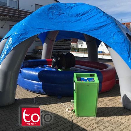 Aufblasbares Rodeogerät mit mechanischem Bullen unter einem blauen Eventzelt, aufgestellt auf einem gepflasterten Platz im Freien.