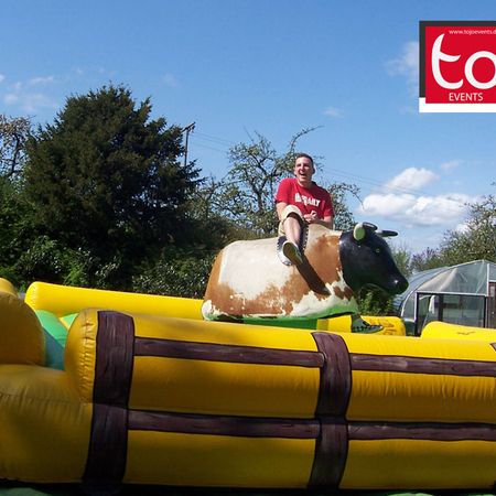 Kind reitet auf einem mechanischen Bullen auf einer gelb-grünen Matte in einer Gartenlandschaft bei Tageslicht.