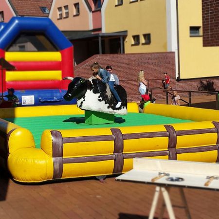 Kinder und Erwachsene beim Rodeo-Reiten auf einem mechanischen Bullen auf einer gelb-grünen Matte, mit bunter Hüpfburg im Hintergrund.