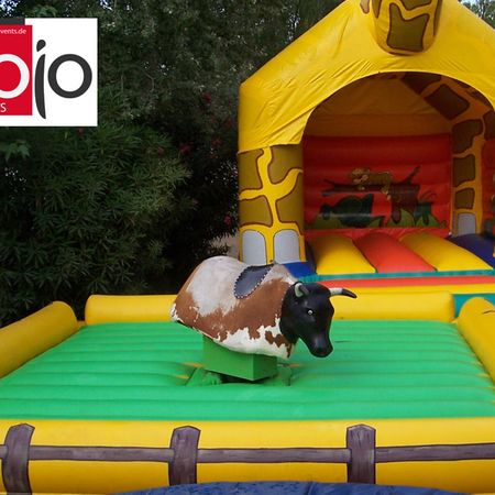Aufblasbarer Rodeo-Bulle vor einer gelben Hüpfburg mit buntem Eingang, aufgestellt auf einem Outdoor-Spielgelände.