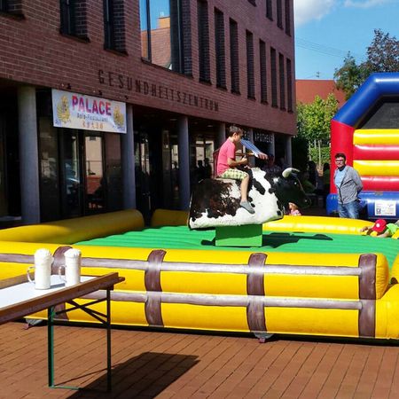 Rodeo-Attraktion mit aufblasbarem gelbem Schutzrahmen, mechanischem Bullen und Menschen in einem belebten Außenbereich.