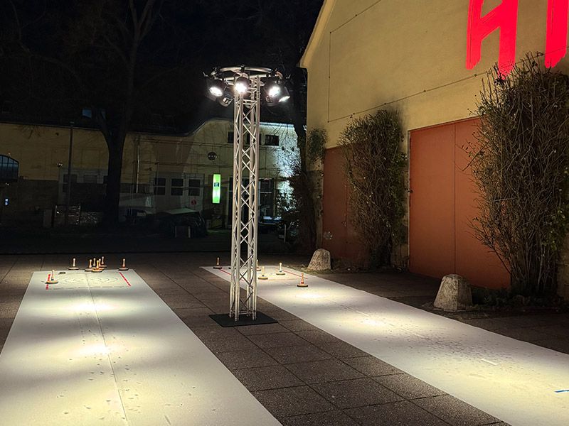 Nachtaufnahme einer beleuchteten mobilen Eisstockbahn vor einem Gebäude, bereit für ein Eventspiel.