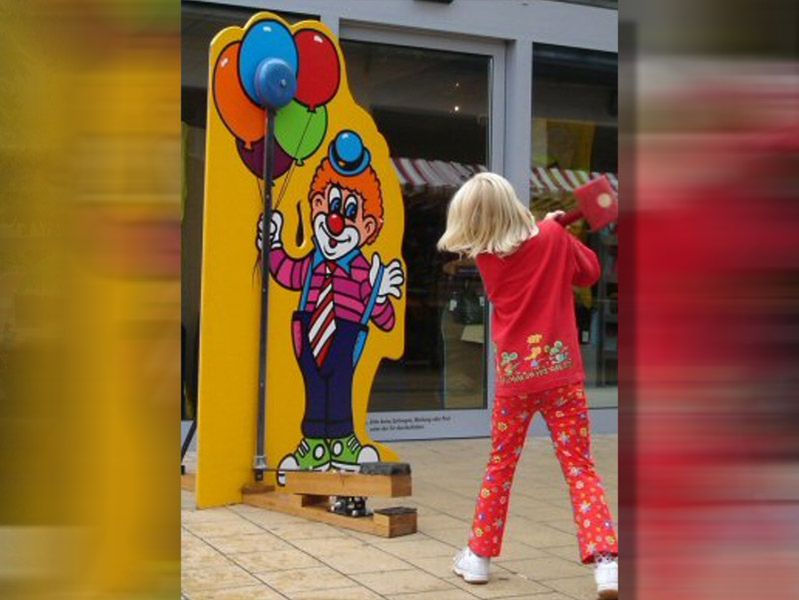 Kind mit blondem Haar und rotem Outfit schlägt mit einem kleinen Hammer auf ein Jahrmarktspiel für Kinder. Das Spiel zeigt eine bunte Clownfigur mit Luftballons auf gelbem Hintergrund, bei dem ein Gewicht nach oben gegen eine Glocke geschlagen werden kann.